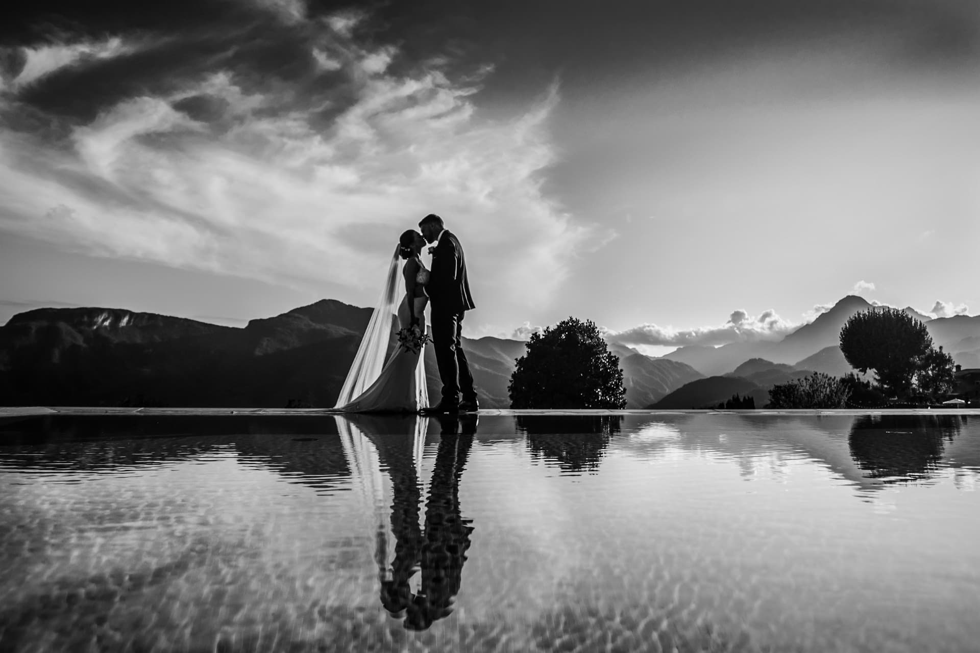 Sposi che si baciano al tramonto riflessi in una piscina, con le montagne sullo sfondo